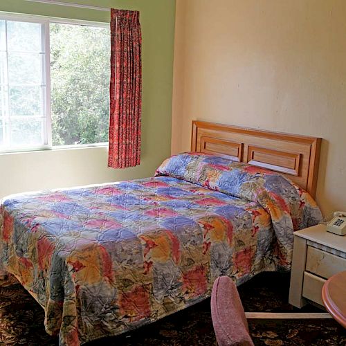 A modest bedroom with a single bed, colorful quilt, side table, telephone, TV, fan, and a window with floral curtains is shown in the image.