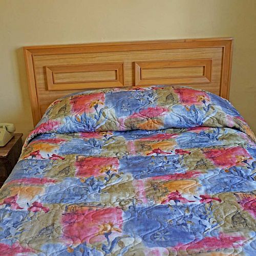 The image shows a bed with a colorful quilt. A wooden headboard is visible, and a bedside table with a phone and remote is to the left.