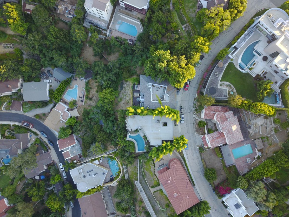 An aerial view of a residential neighborhood with several houses, green trees, and swimming pools, intersected by curving roads.