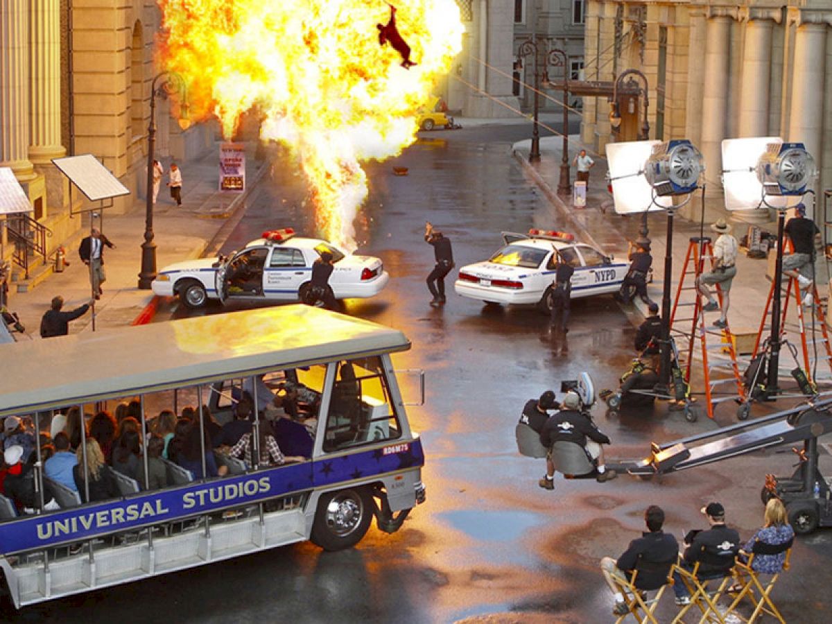 A studio tour bus passes a film set with an explosion, police cars, and crew. Cameras and lighting are visible, capturing the dramatic scene.