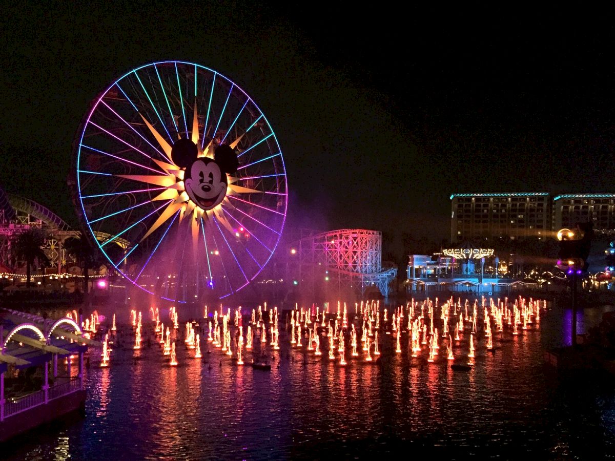 The image shows a vibrant nighttime scene at a theme park with a large Ferris wheel featuring a recognizable character, illuminated fountains, and festive lights.