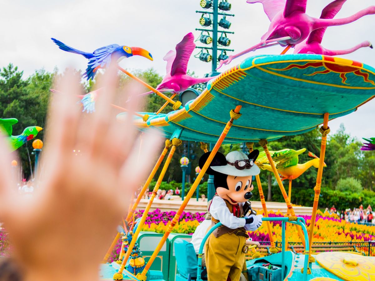 A person dressed as a popular cartoon character is part of a colorful outdoor parade, with an out-of-focus hand waving in the foreground.