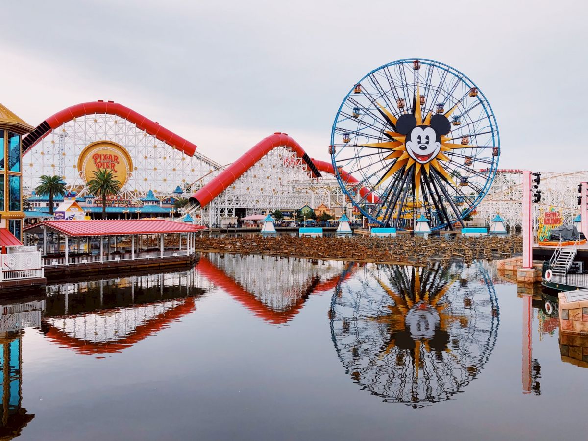 The image shows a theme park with a Ferris wheel featuring a cartoon character's face, roller coasters, and a water body, all reflecting in the water.