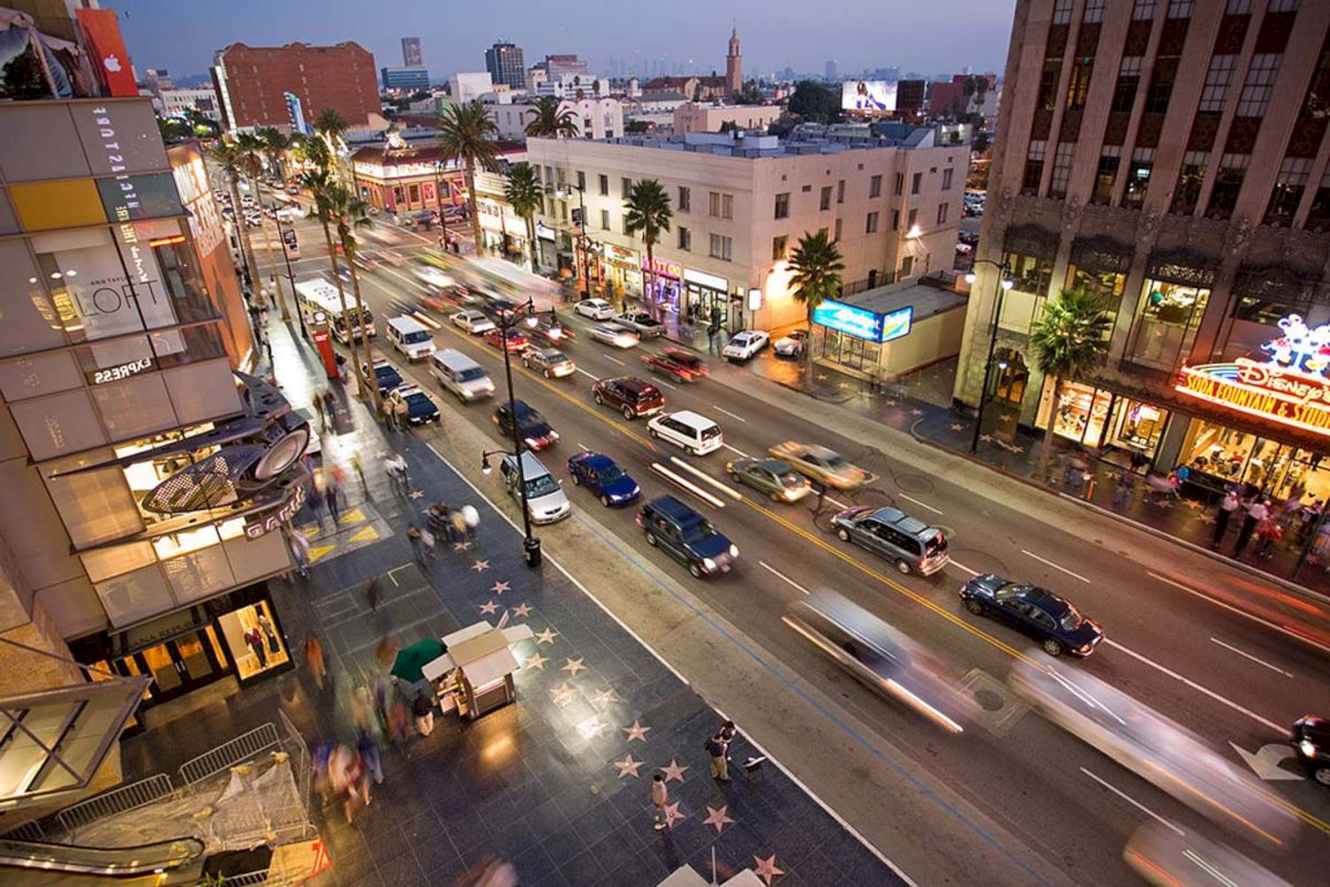 A wide-angle view of a bustling street with cars and people, lined with buildings and palm trees, and a star-studded sidewalk.