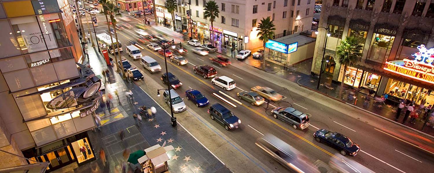 A nighttime view of a busy street with cars and pedestrians, showing several buildings and a sidewalk with stars.