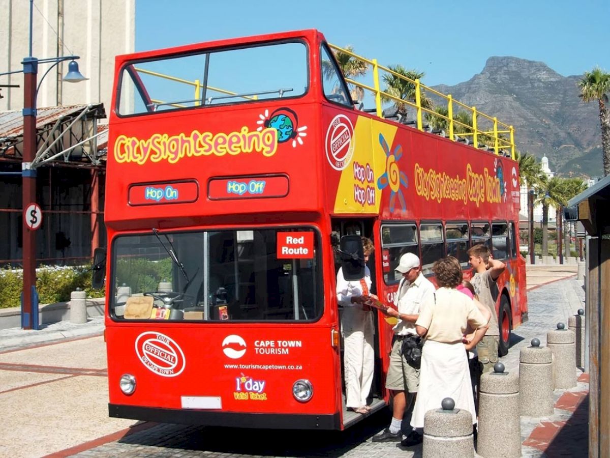 A red double-decker "City Sightseeing" tour bus is parked, with people boarding or interacting near the entrance, set against a scenic background.