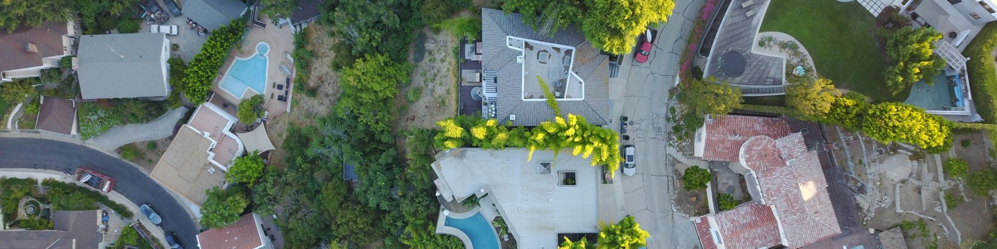 This image shows an aerial view of a neighborhood with houses, backyards, and pools, surrounded by trees and roads, forming a residential area.