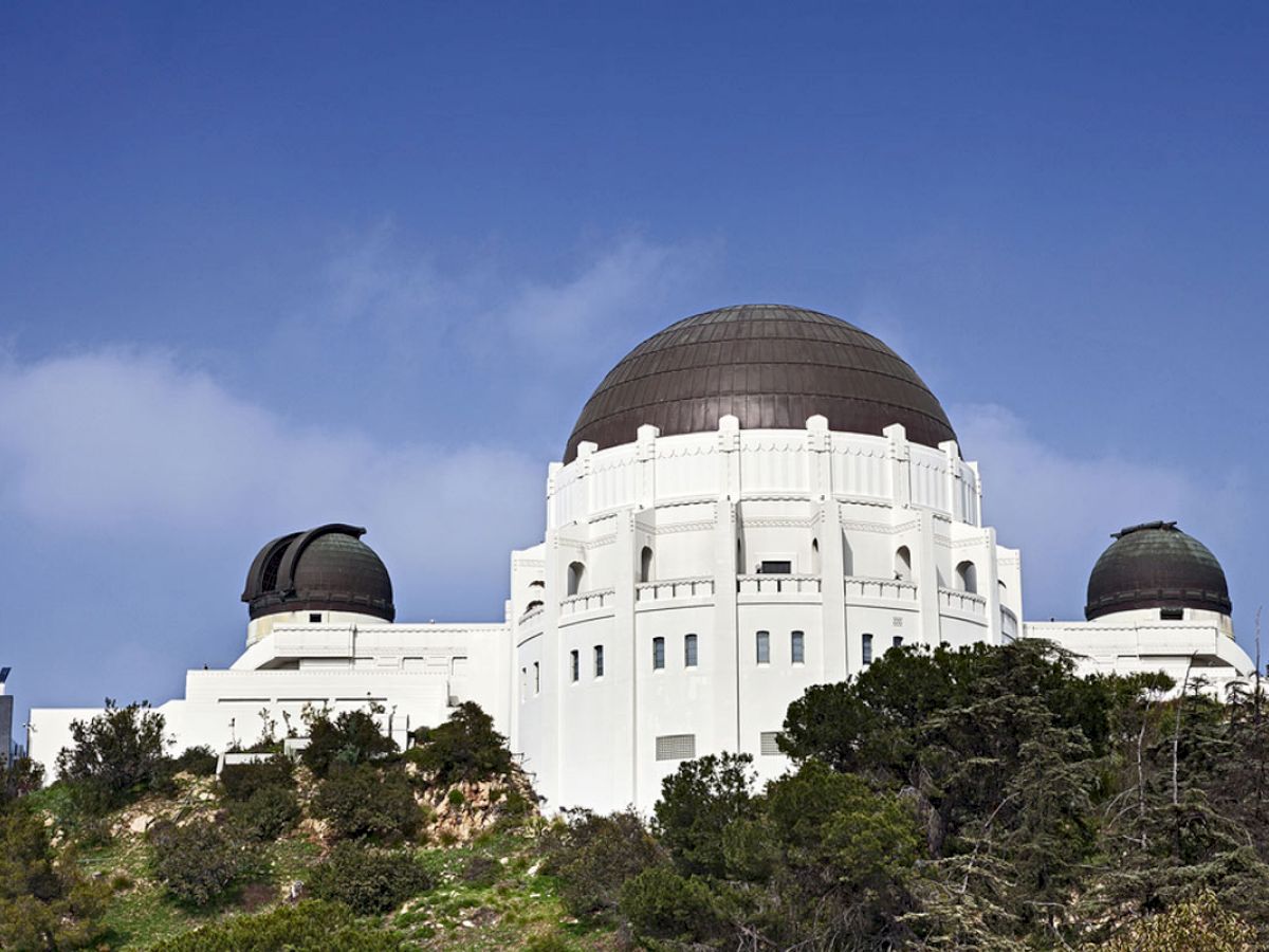 The image shows a white observatory with three domes located on a hill with greenery, under a blue sky.