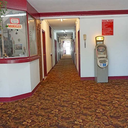 A hallway with carpeted flooring, a manager's office, a vending machine, plants, and a stairway to the left. There's a brochure stand on the right.