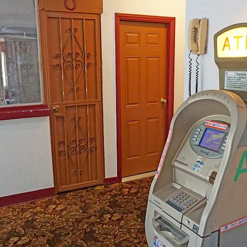 An indoor setting with an ATM machine, a wooden door, and a counter. A wall phone is mounted nearby. Carpeting and a clock are also visible.