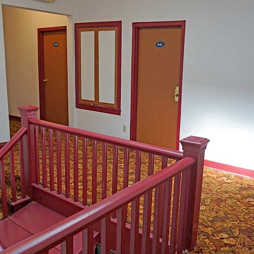 The image shows an interior hallway with a crimson-red staircase and carpeted floor leading to doors with room numbers, likely in a hotel or apartment.