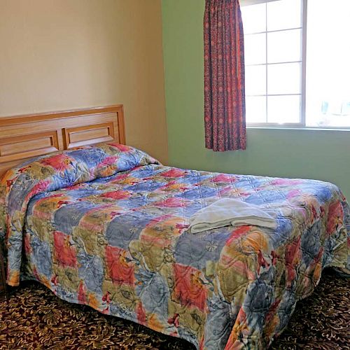 A cozy room with a bed covered in a colorful quilt, a side table with a phone, curtains, a window, and a TV on a wooden dresser.