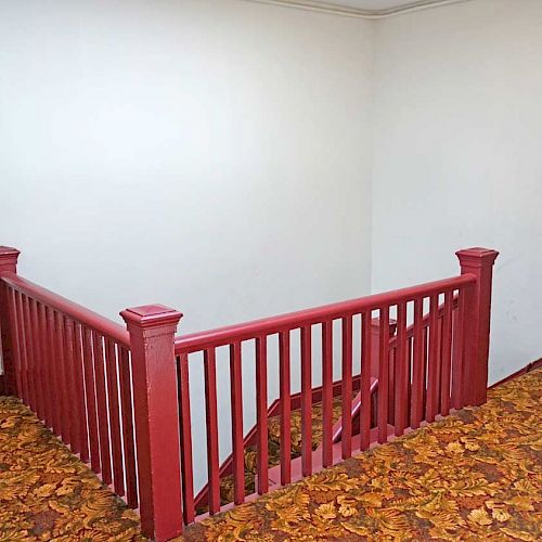 A hallway with red trim and orange-patterned carpet, featuring a staircase railing in the center and doorways on the left and right walls.