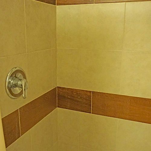 This image shows a tiled shower area with beige and brown tiles, featuring a showerhead and a control knob on the wall.