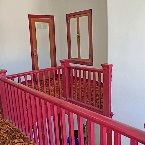 An indoor hallway features a red staircase railing, carpeted floor, white walls, and a wooden door with a window.