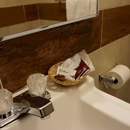 A bathroom sink with dual faucet knobs, a basket of toiletries, a partially visible mirror and towel, and a toilet paper holder.