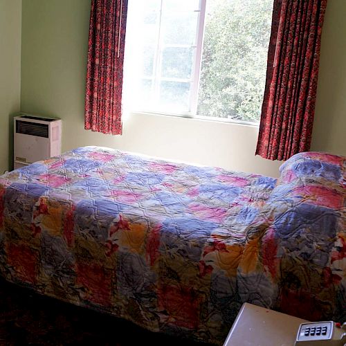 This image shows a bedroom with a bed covered in a colorful quilt, a nightstand with a telephone and lamp, and a window with red curtains.