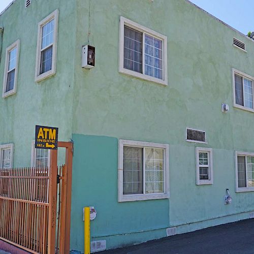 This image shows a green, two-story building with a hotel sign and an ATM machine near the entrance, with some windows and a fence.