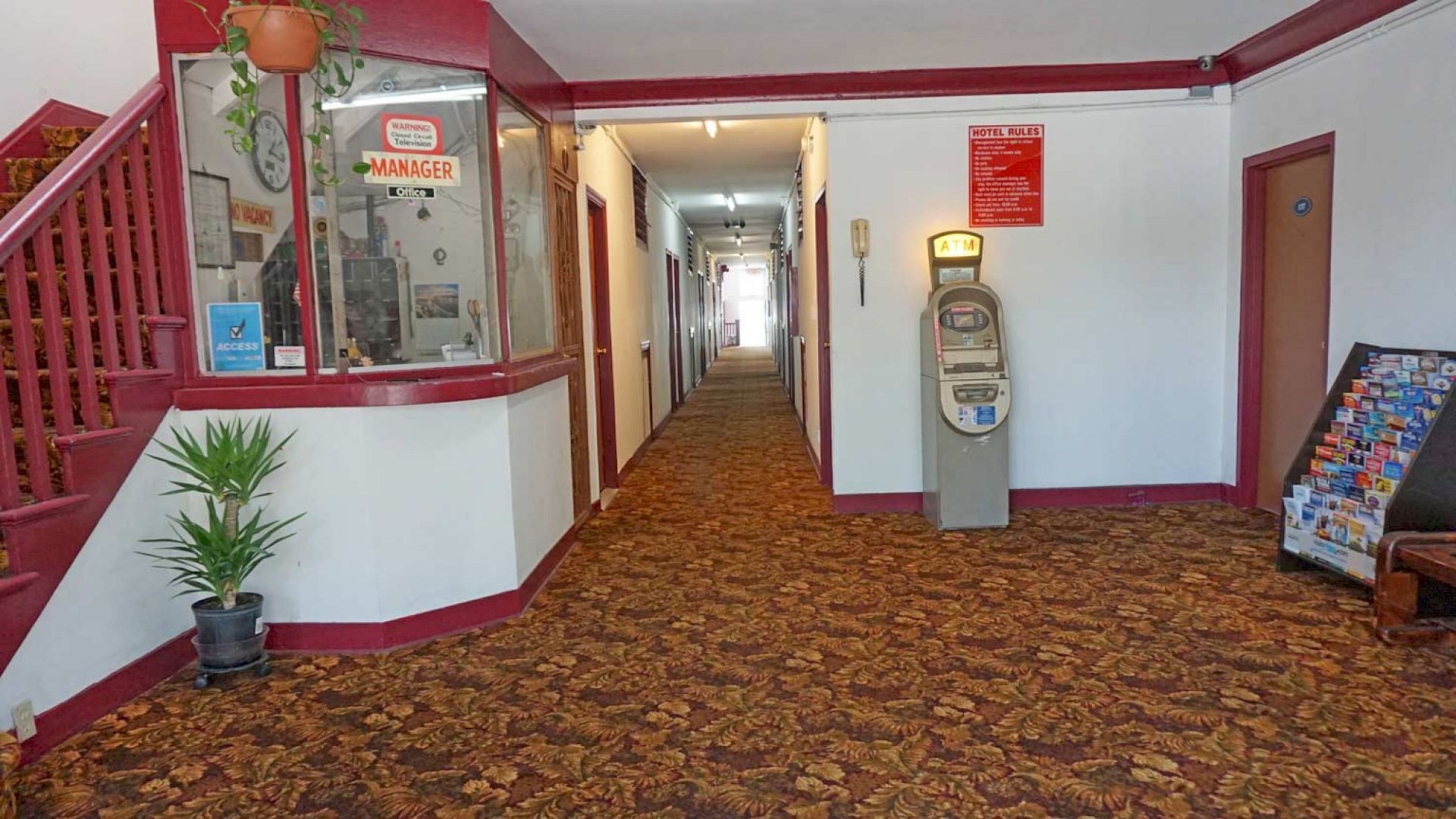 A hallway with carpet, a manager's office, an antique scale, a potted plant, and a brochure stand, with doors along the hallway.