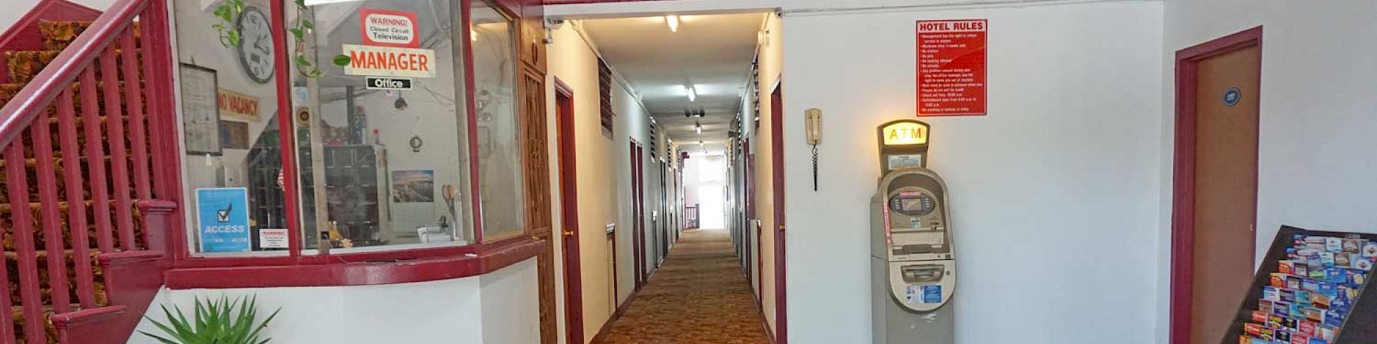 A hallway with carpet, a manager's office, an antique scale, a potted plant, and a brochure stand, with doors along the hallway.