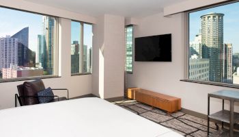 A modern bedroom with large windows showing a cityscape, featuring a bed, chair, TV, and wooden bench, ending the sentence.