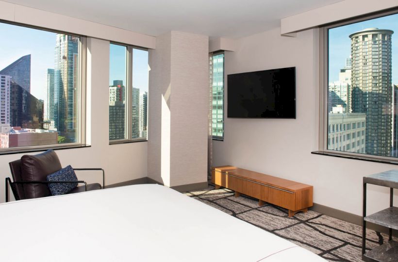 A modern bedroom with large windows showing a cityscape, featuring a bed, chair, TV, and wooden bench, ending the sentence.
