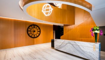 A modern interior with a reception desk, a large wall clock, decorative lighting, wooden walls, and a vase of flowers on the counter.