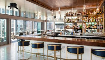 A modern bar with blue-cushioned stools, a well-stocked liquor display, pendant lights, and large windows with a view of the cityscape outside.