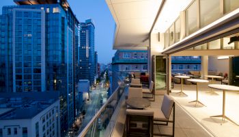 The image shows a modern cityscape from a high-rise balcony at dusk, with glass buildings, outdoor seating, and illuminated interiors.