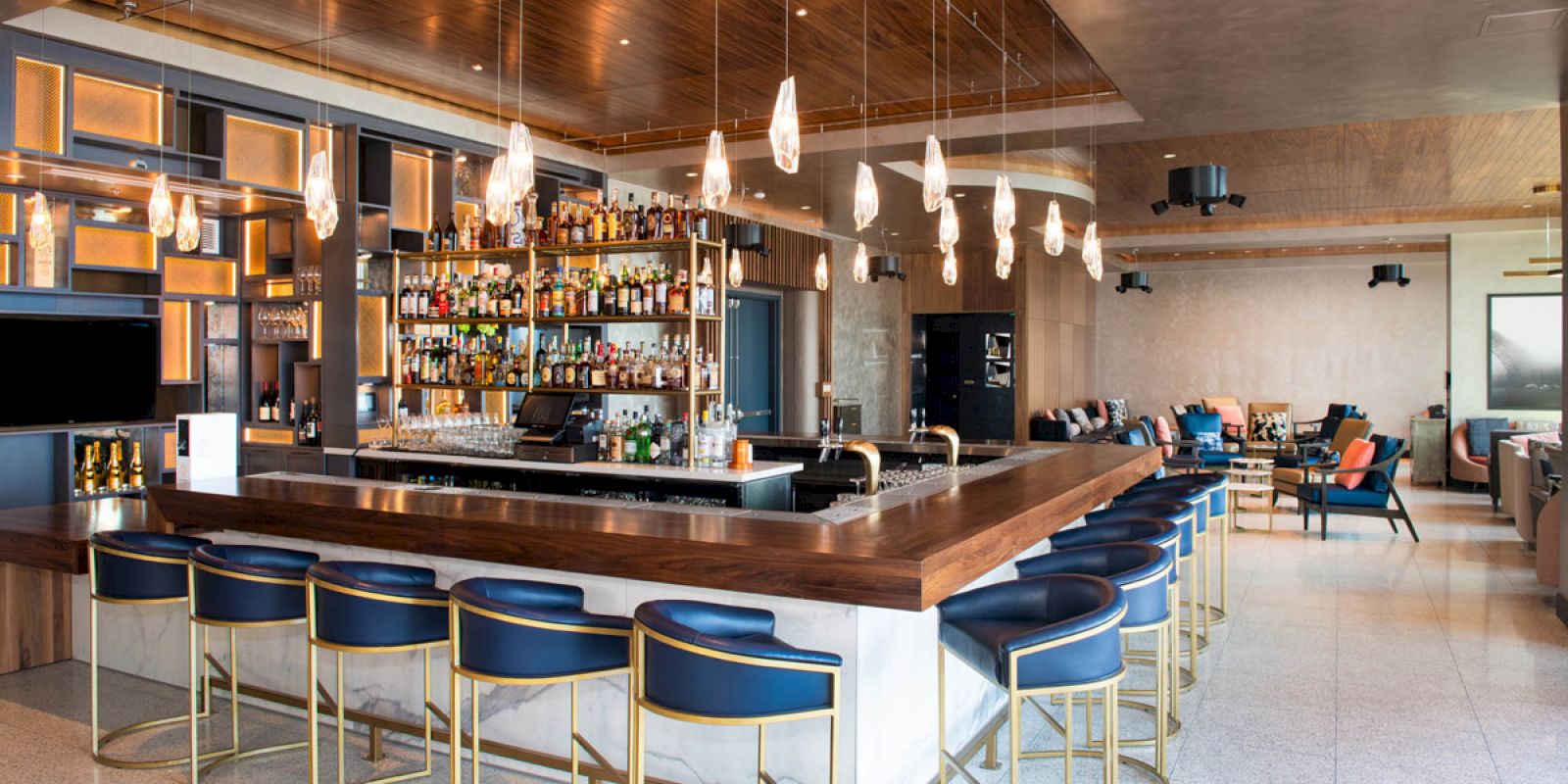 A modern bar with blue stools, a well-stocked bar, hanging lights, and a seating area with couches and chairs.