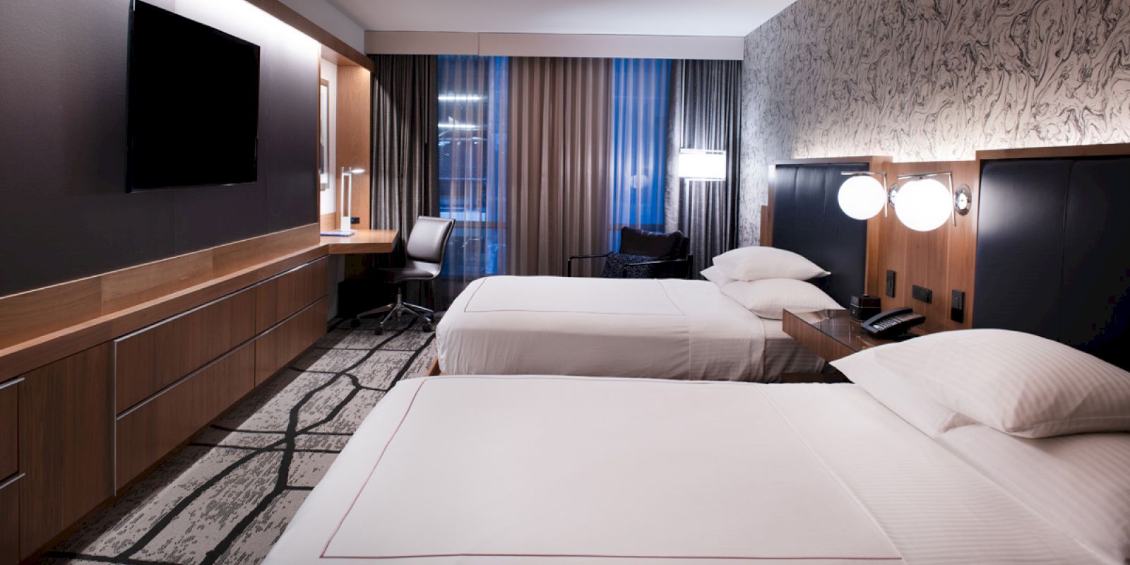 This image shows a hotel room with two double beds, a mounted TV, a desk with a chair, modern decor, and ambient lighting.