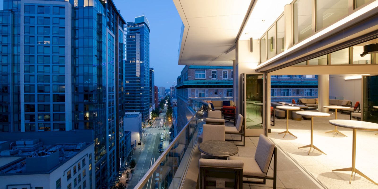 A modern balcony with seating overlooks a city street lined with tall buildings at dusk, capturing the urban skyline and lights.