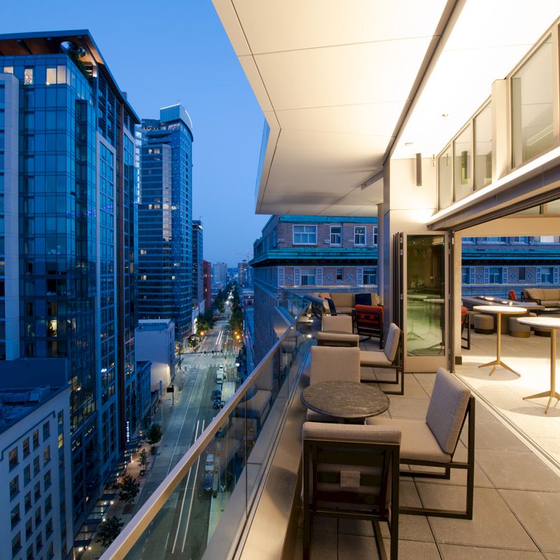 A modern cityscape at dusk viewed from a balcony with tables and chairs facing the skyline, featuring tall glass buildings and a lit street below.