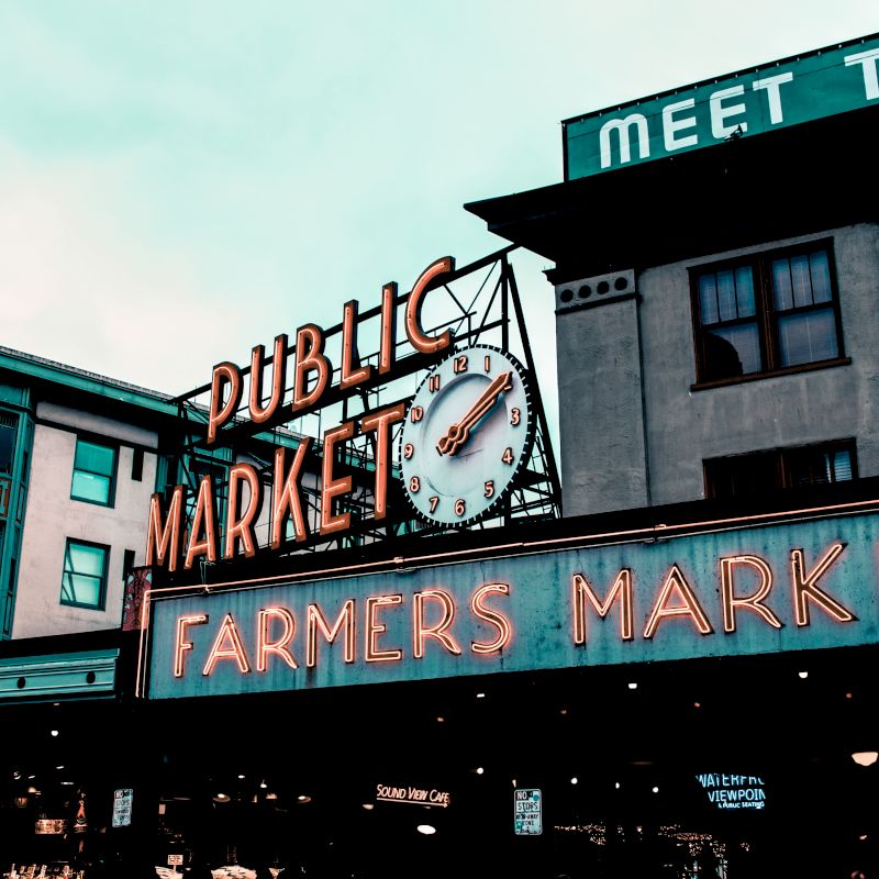 This image shows the iconic neon signs for "Public Market" and "Farmers Market" at Pike Place Market in Seattle. The background includes buildings.