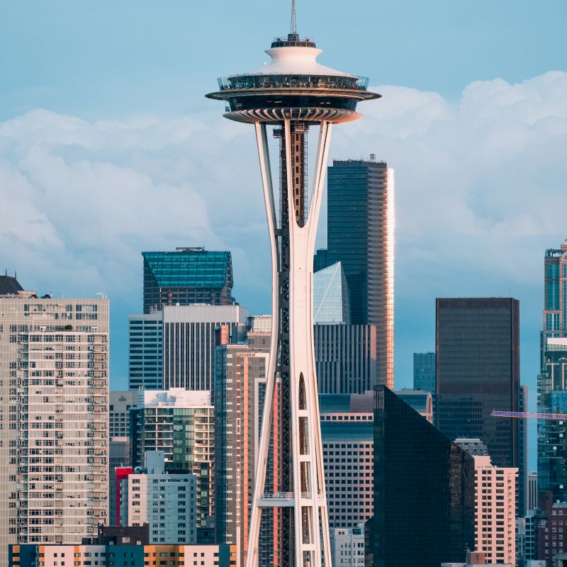 The image shows a city skyline with a prominent observation tower. Skyscrapers surround the tower, and the sky is partially cloudy.