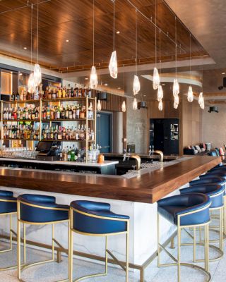 A modern bar setting with a wooden countertop, blue bar stools, hanging lights, and a fully stocked bar in the background with seating areas.