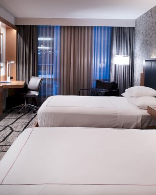 The image shows a modern hotel room with two neatly made beds, a TV, a desk and chair, and a window with curtains, reflecting a cozy ambiance.