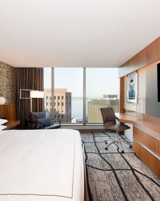 A modern hotel room with a large bed, a flat-screen TV, a desk with a chair, and a large window offering a view of buildings and water.