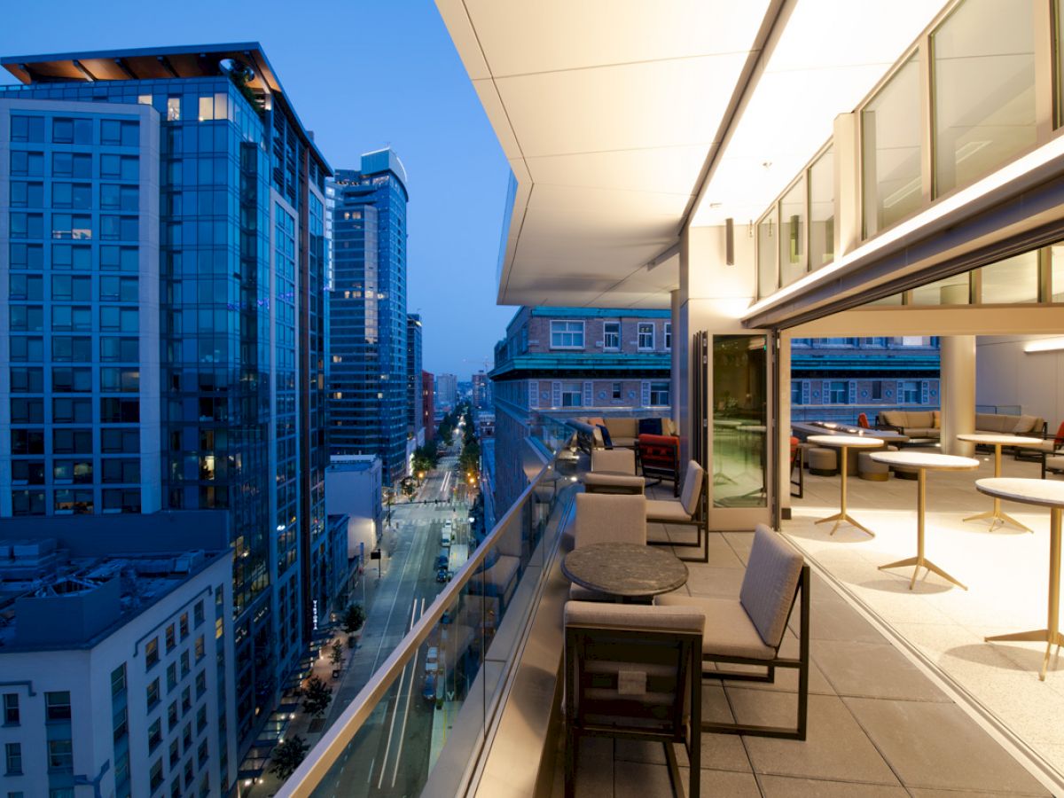 An urban balcony scene at twilight, featuring modern furnishings and a view of tall buildings and a city street below, captured in the evening.