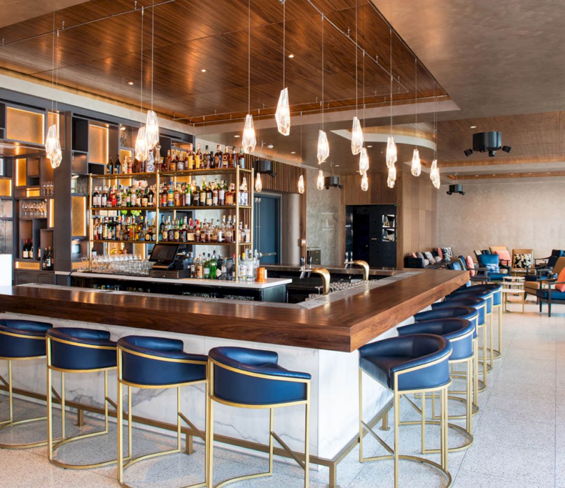 A wide view of a stylish bar with a long wooden counter, blue stools, and gold accents.