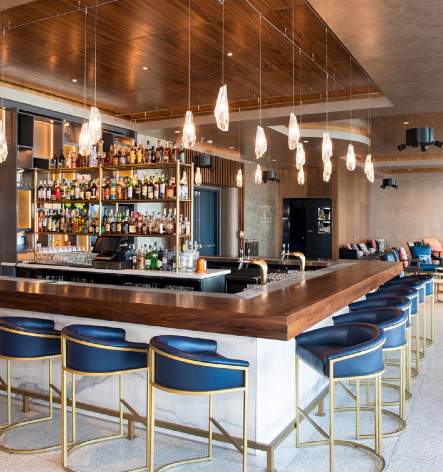 The image shows a modern bar with a wooden counter, blue barstools, a well-stocked liquor shelf, pendant lights, and a spacious seating area.