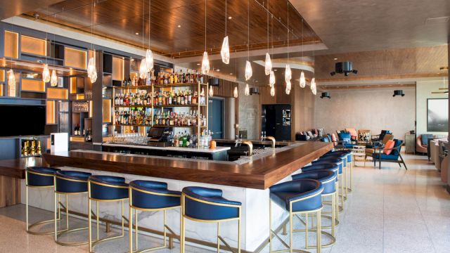A modern bar with a long wooden counter, blue stools with gold frames, and a well-stocked liquor shelf.