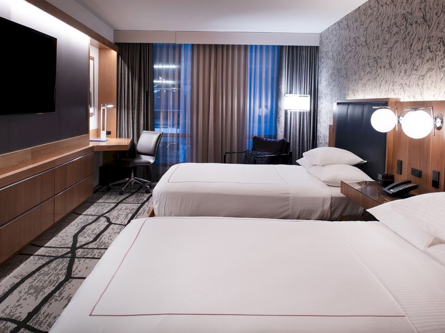 The image shows a modern hotel room with two neatly made double beds, a flat-screen TV, a desk with a chair, and stylish decor with patterned wallpaper.