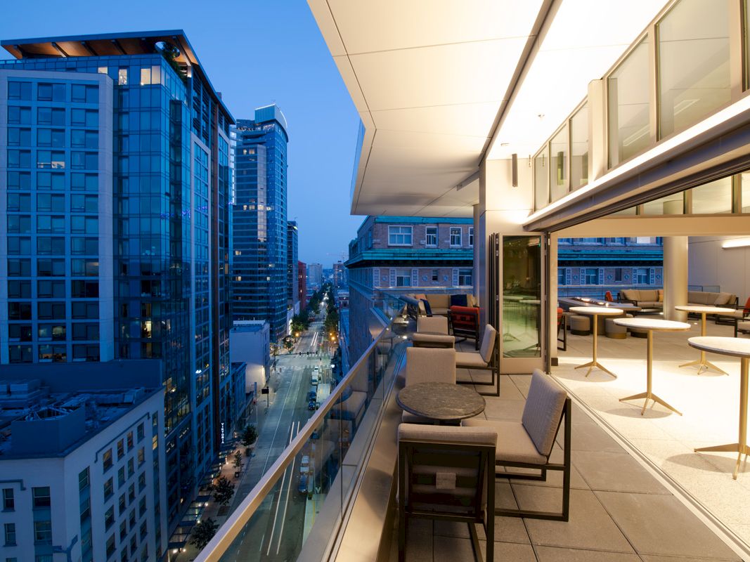 A modern high-rise balcony at dusk overlooks a cityscape with tall buildings and a street below, featuring outdoor seating and a covered area.