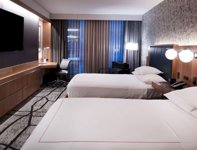 This image shows a modern hotel room with two neatly made beds, a desk and chair, a large TV, and floor-to-ceiling windows with curtains.