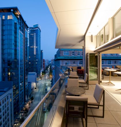 A modern urban balcony overlooking city skyscrapers at dusk, with seating and tables set up for relaxation or socializing.