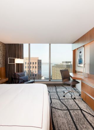 A modern hotel room with a large bed, a desk, a chair, a flat-screen TV, and a window offering a city view with tall buildings in the background.