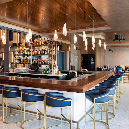 A modern bar with blue chairs, a variety of bottles on shelves, pendant lights, and a seating area in the background.