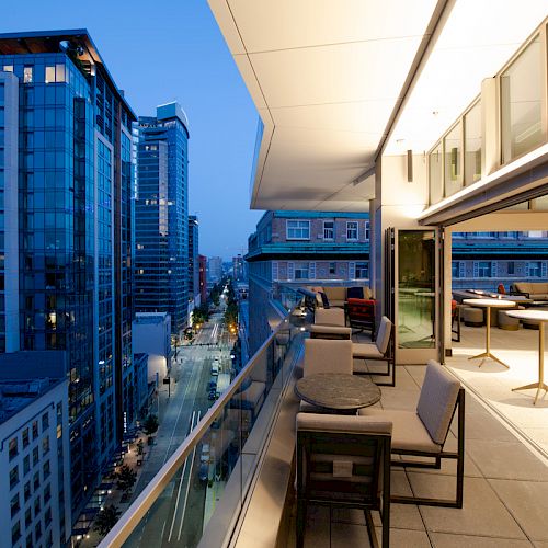 A modern cityscape at dusk with tall buildings, viewed from a high balcony with seating and tables, alongside a brightly lit indoor space.
