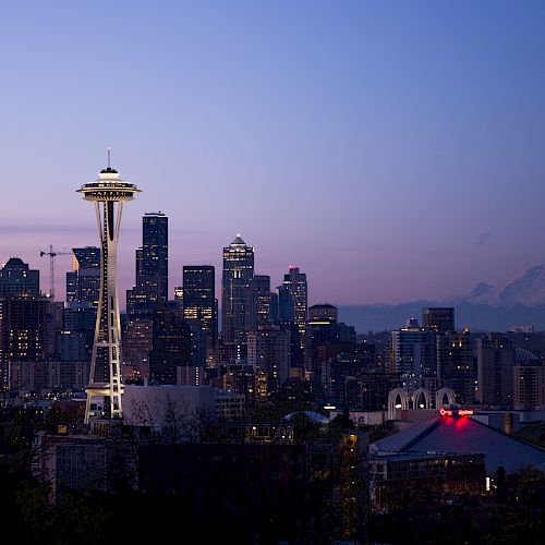 A skyline view of a city at dusk, featuring a prominent tower, numerous skyscrapers, and a distant mountain under a twilight sky.